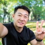 Close-up photo. Selfie of a young Asian male athlete. He works out, runs, does sports in the park, holds the phone, smiles at the camera, shows victory with his fingers.